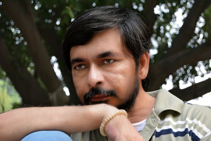 Man with a beard resting his chin on his hand outdoors beside a leafy tree in a calm reflective pose